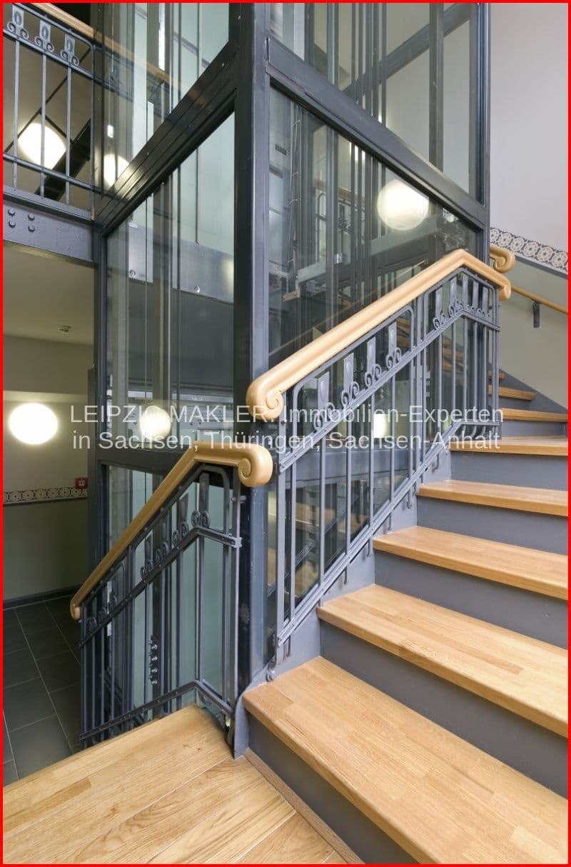 Treppenhaus mit Holzstufen, grauer Wangenverkleidung, Holzhandlauf und Metallgeländer, Aufzug im Hintergrund
