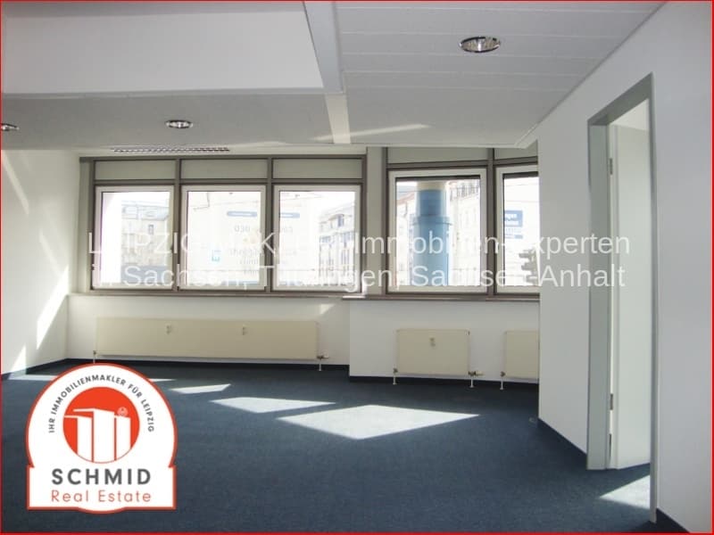 Leerer Büroraum mit Fensterfront, Deckenleuchten, dunklem Bodenbelag, Heizkörpern und Tür zu Nebenraum