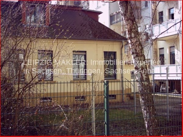 Gelbes Einfamilienhaus mit Satteldach und Metallzaun, dahinter mehrstöckiges Wohngebäude, kahl Birken und Äste davor