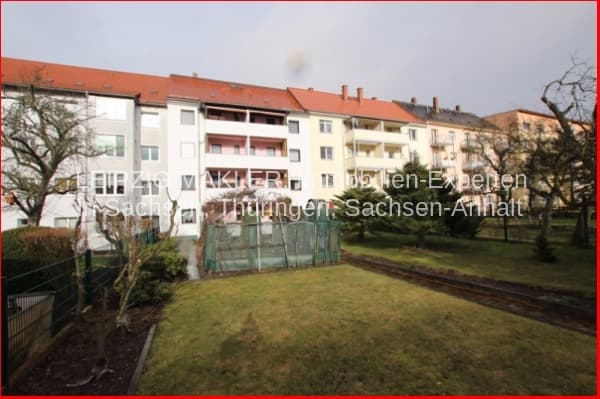 Mehrfamilienhaus mit Flachdach und Ziegeldachgiebel, vier Geschosse, Balkonreihen, Grünfläche davor, Zaun und Bäume