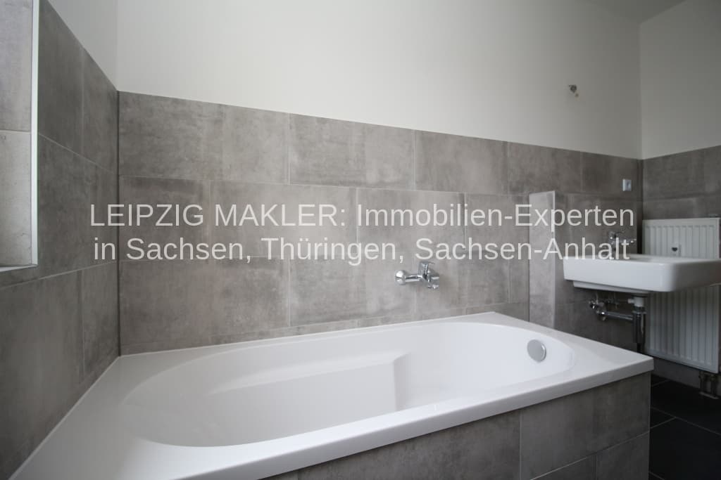 Badezimmer mit weißer Badewanne, grauen Feinsteinzeugfliesen an den Wänden, weißem Waschbecken und Heizkörper