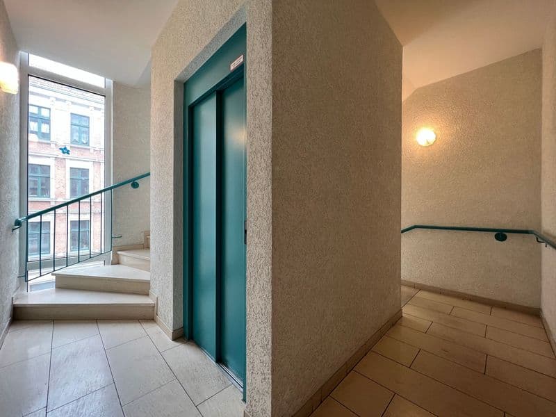 Treppenhaus mit Aufzug, Betonpfeiler, Fliesenbelag im Eingangsbereich, Holzbelag im Flur, grüne Aufzugstür