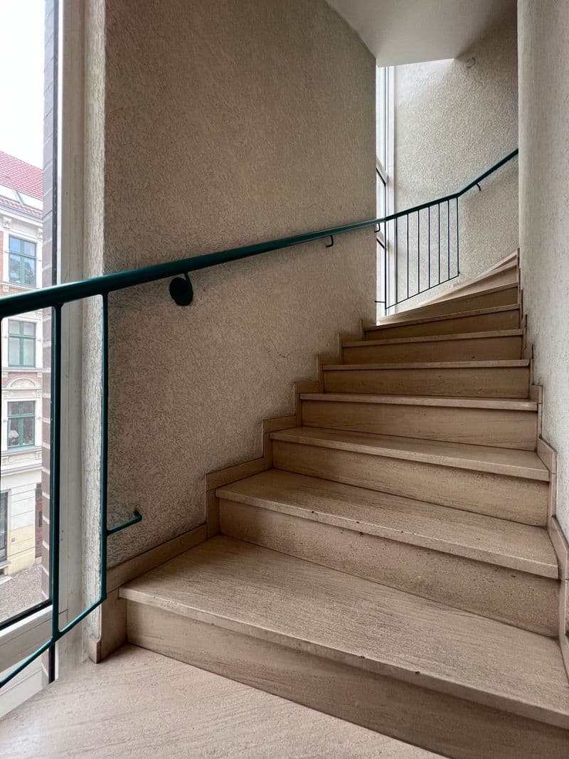 Trepenhaus mit Holzstufen, beige Wänden, schwarzem Handlauf und Fenster mit Blick auf Nachbargebäude