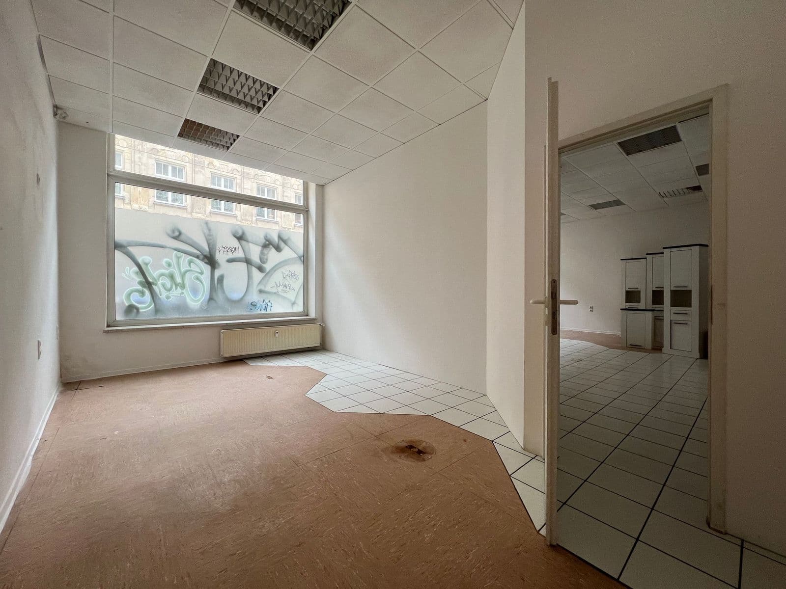 Leerer Ladenraum mit Fensterfront, Deckenleuchten, unterschiedliche Bodenbeläge, Tür zu Nebenraum