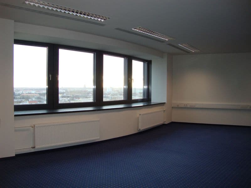 Leerer Bueroraum mit vier schwarzen Fensterflügeln, weißer Wand, Heizkörper und blauem Teppichboden, Blick auf Stadt