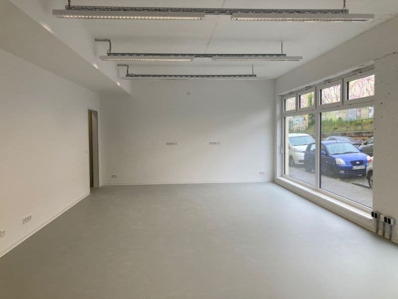 Leerer Gewerberaum mit Betonboden, weiße Wände, Deckenleuchten, große Glasfront zur Straße