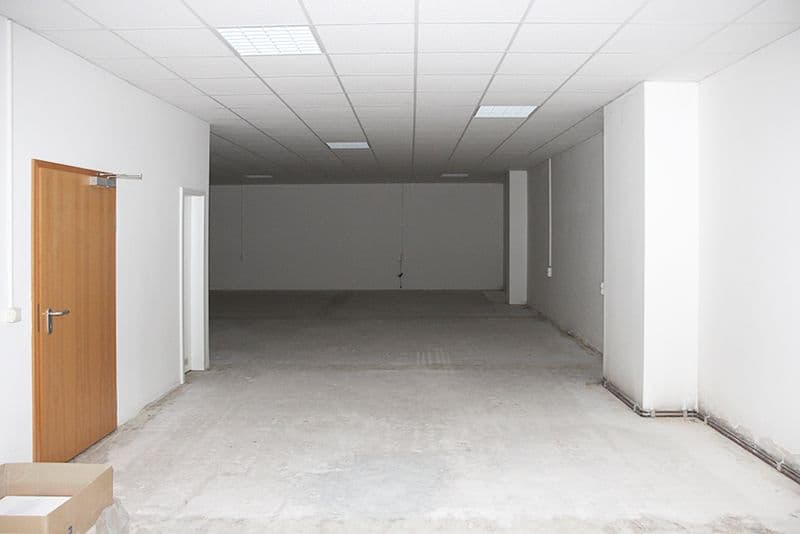 Leerer Gewerberaum mit Betonboden, abgehängter Decke mit LED-Einbauleuchten, weiße Wände, Holztür links