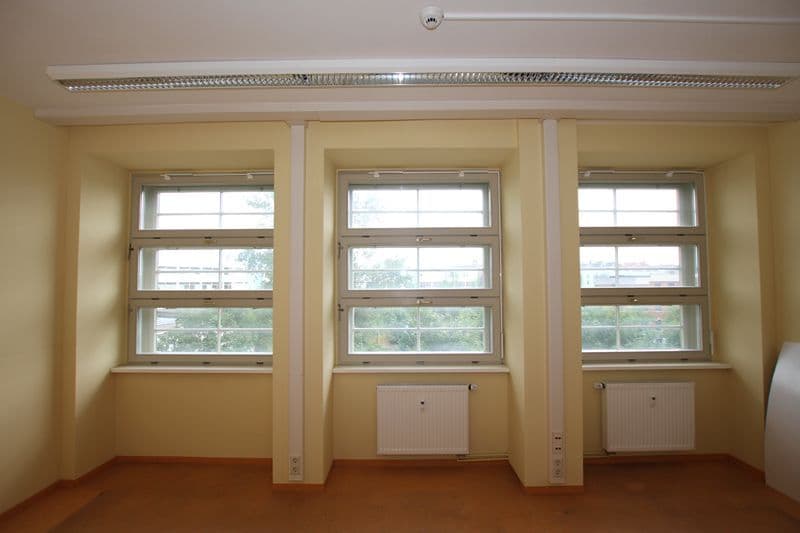 Leerer Büroraum mit drei großen Fensterblöcken, gelbbeiger Wandfarbe, Terrakotta-Estrich, Heizkörper und LED-Deckenleuchte
