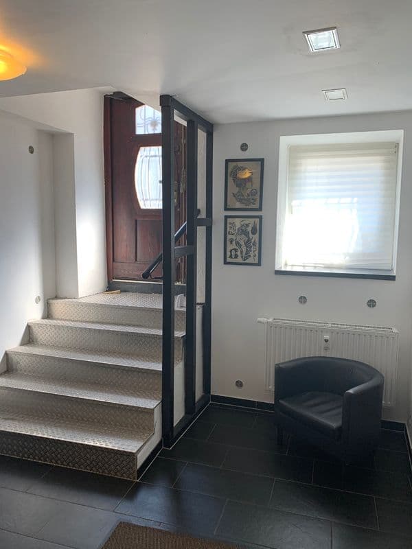 Treppenhaus mit dunklem Fliesenboden, stahlverkleideter Treppe mit gemustertem Belag, schwarzer Metalltür