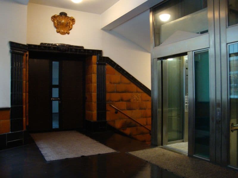 Hausflur mit schwarzer Holztür, Treppe mit orange-brauner Wange, moderner Aufzug mit Glastüren