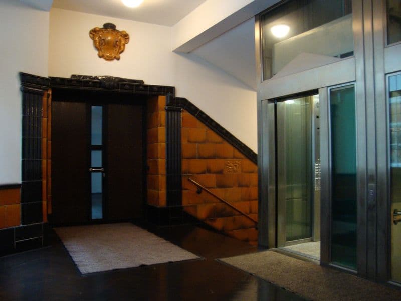 Hausflur mit Aufzug, schwarze Eingangstür mit Glasausschnitten, Treppe mit orangefarbenen Stufen, Petersstraße in Leipzig