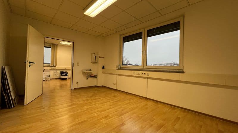Leerer Büroraum mit Holzparkett, heller Wand- und Deckenverkleidung, großem Fenster und offener Tür zu Nebenraum