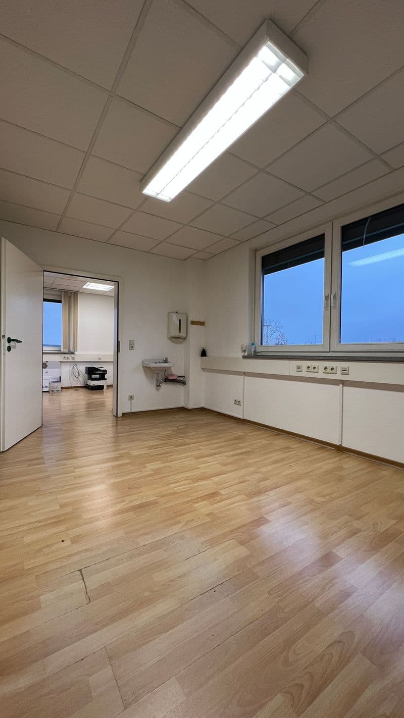 Leerer Büroraum mit Parkettboden, Deckenleuchte, großen Fenstern und offener Tür zum angrenzenden Raum