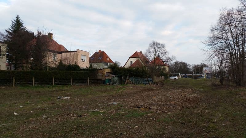 Freie Fläche mit Holzzaun, dahinter Wohnhäuser mit Ziegeldächern und Gartenbereiche, Prager Straße in Leipzig