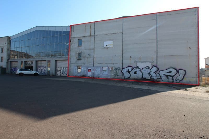 Großflächige Gewerbehalle mit rotem Stahlrahmen, vergraute Betonwand, wenige Fenster, Graffiti an der Seitenwand