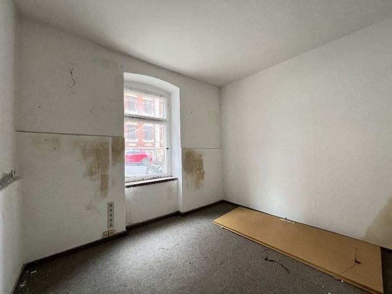 Leerer Raum mit Betonboden, hellen Wänden mit Feuchtigkeitsschäden, Fenster zur Straße, Steckdosen sichtbar