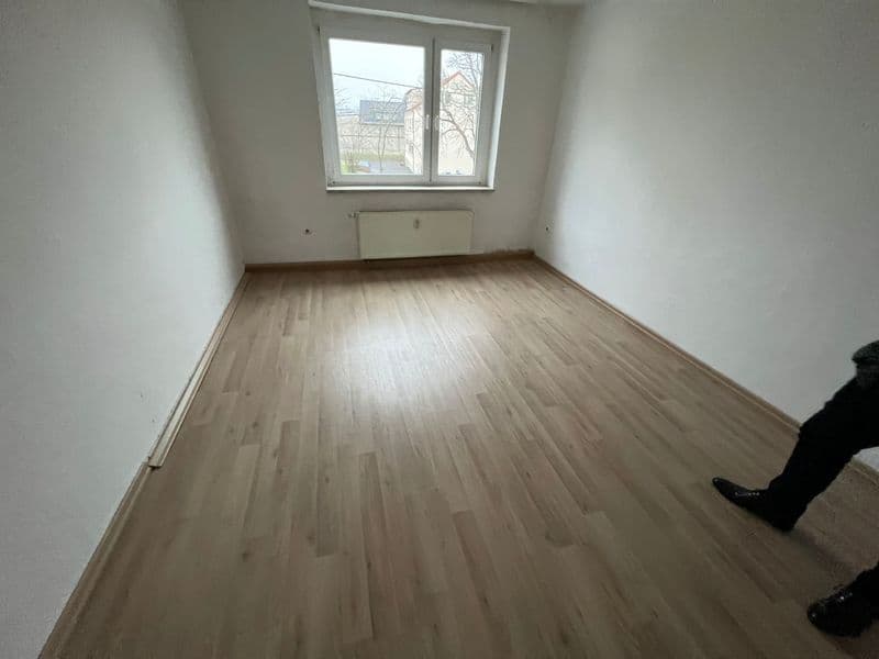 Leerer Wohnraum mit Holzstabparkett, weiße Wände, Fenster mit Blick auf Außenanlage