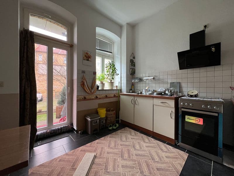 Küche mit weißen Schrankelementen, schwarzem Gasherd, Fliesenwand, Fenster zur Straße und Fliesenboden