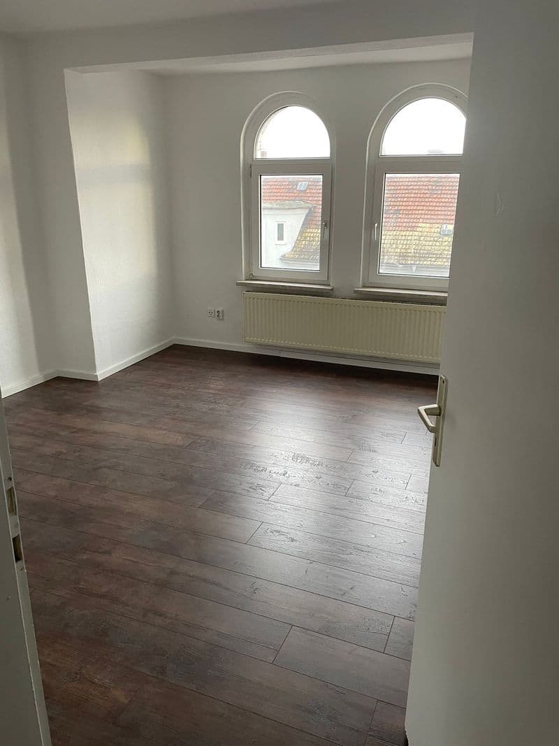 Leerer Raum mit Parkettboden, weiße Wände, zwei Bogenfenster mit Heizkörper, Blick auf Ziegeldächer
