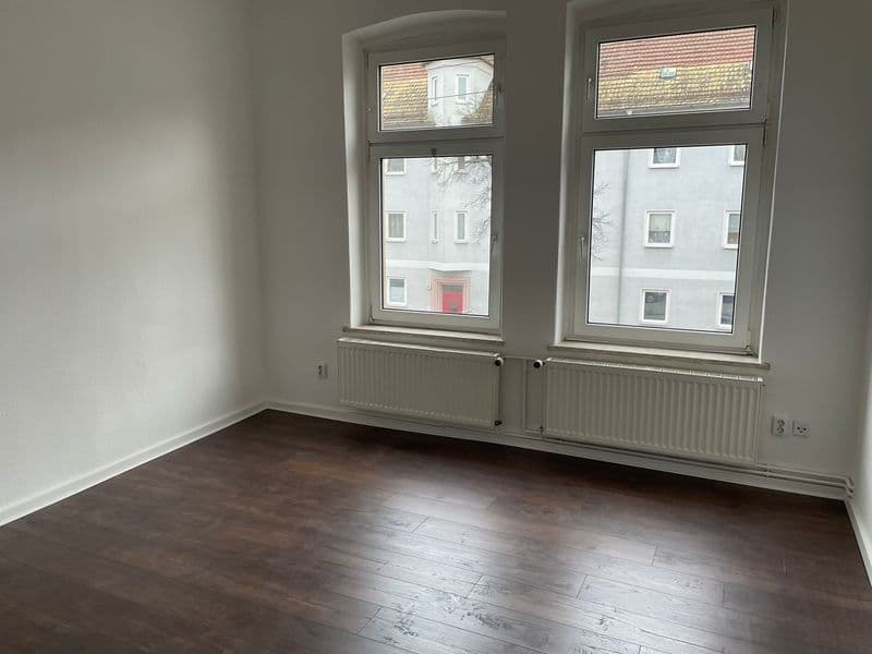 Leeres Zimmer mit Parkettboden, weiße Wände, zwei Fenster mit Heizkörpern, Blick auf Wohnhaus gegenüber