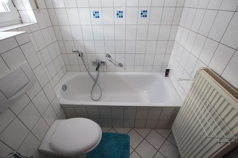 Badezimmer mit weißer Wandfliesenverkleidung, Badewanne mit Duscharmatur, WC und Heizkörper