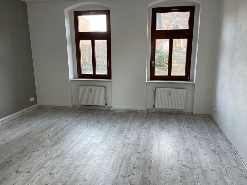 Leeres Zimmer mit hellem Holzlaminatboden, zwei Sprossenfenstern mit dunkelroten Rahmen und Heizkörpern