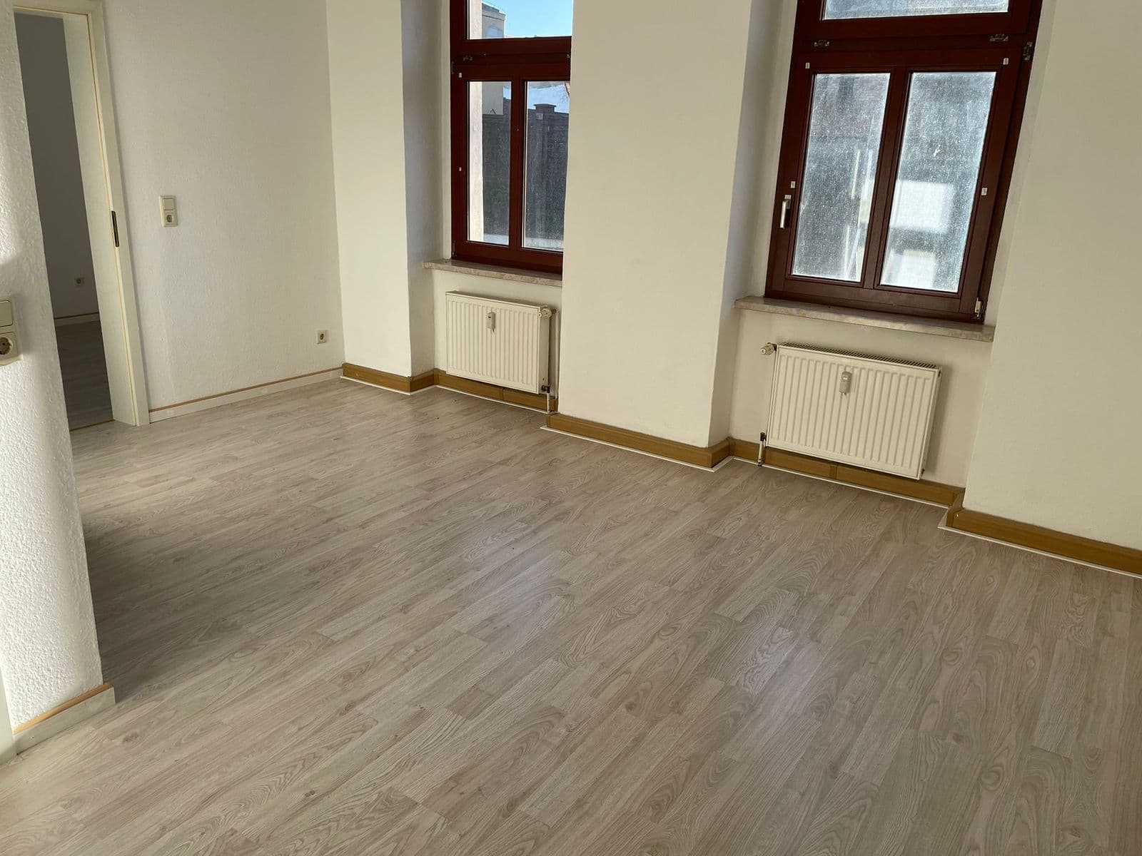 Leerer Wohnraum mit Vinylboden, helle Wände, zwei Fenster mit Holzrahmen, Heizkörper, Sockelleiste aus Holz