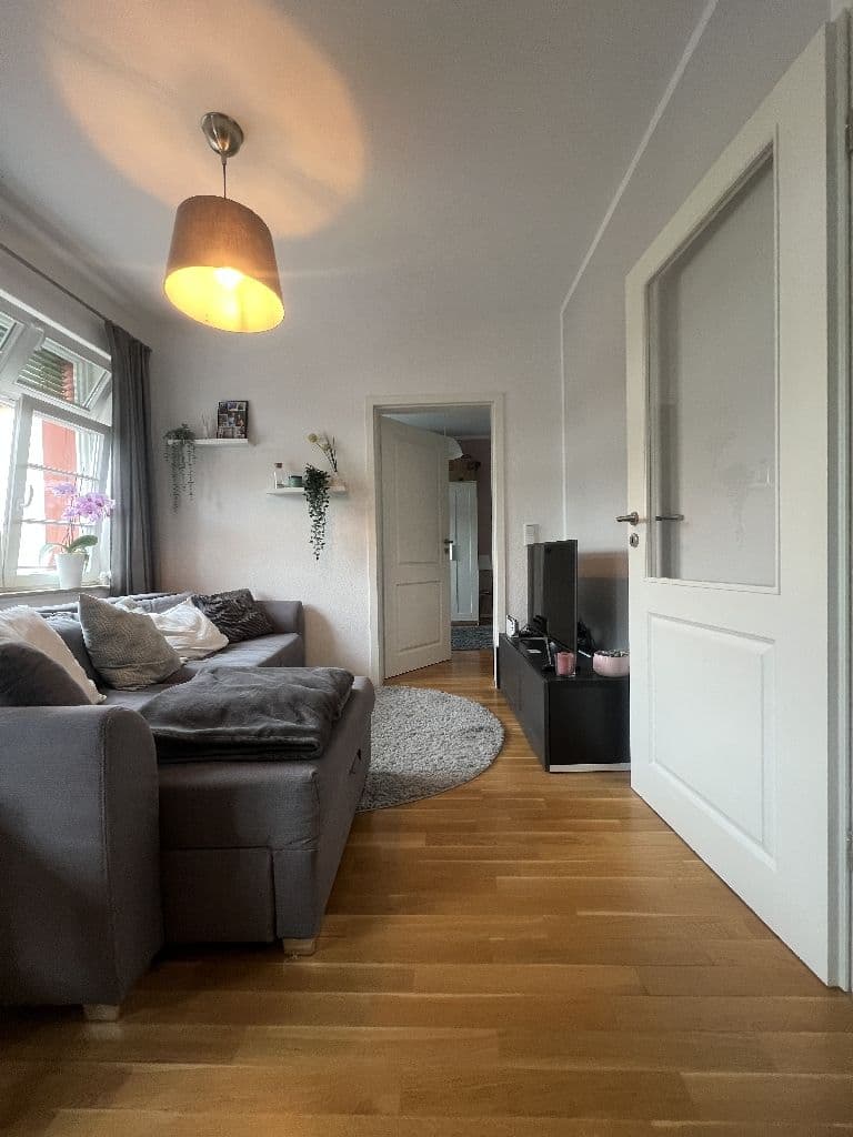 Wohnzimmer mit grauem Polstersofa, Holzparkett, weißen Türen, Fenster mit Gardinen und hängender Holzleuchte
