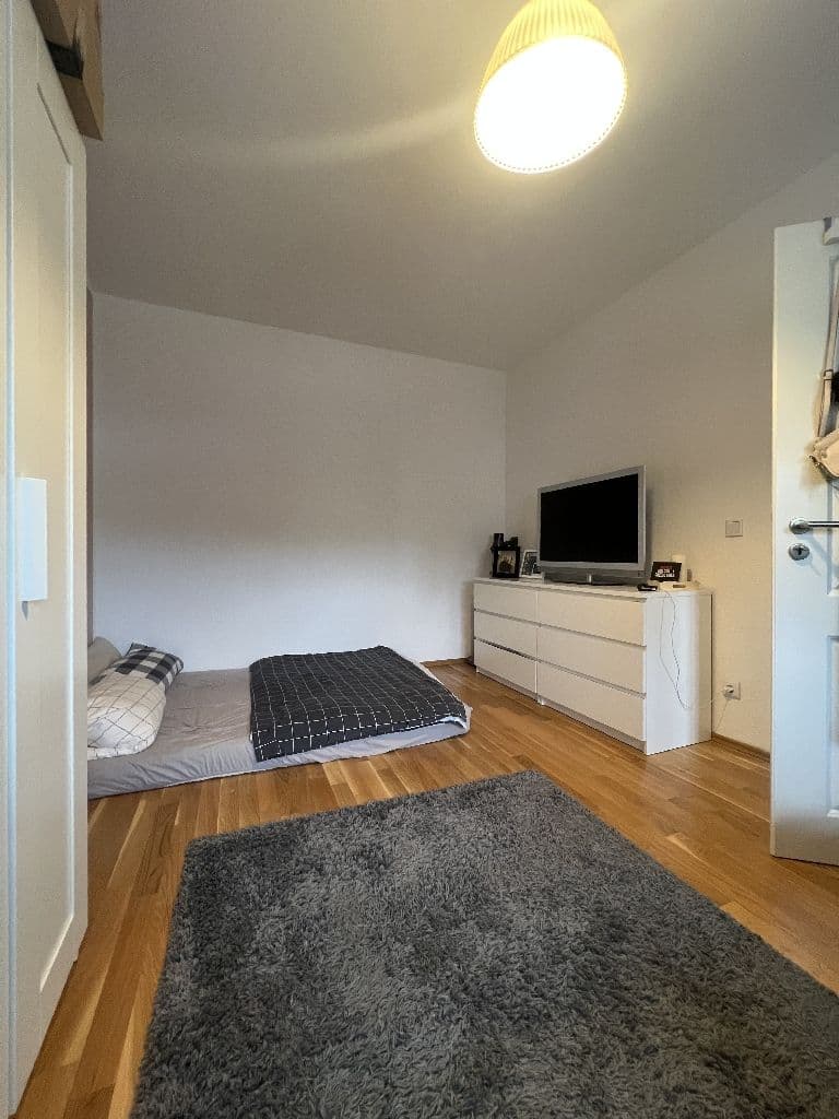 Schlafzimmer mit Parkettboden, Bett, weißer Kommode mit Fernseher und grauem Teppich