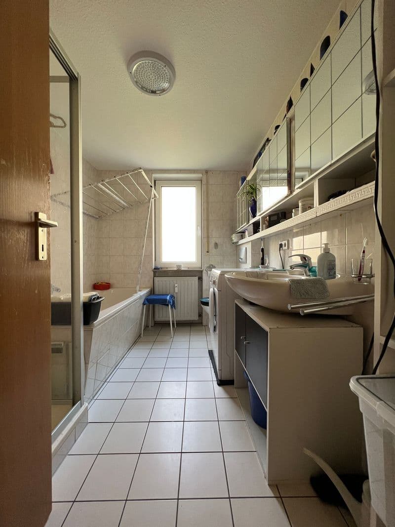 Schmales Badezimmer mit Fliesenboden, Wanne links, Waschbecken rechts mit Spiegelschrank, Fenster in der Mitte