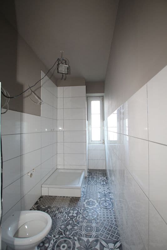 Schmales Badezimmer mit weißen Wandfliesen, Toilette links, Duschbecken Mitte, Tür zum Flur, gemusterter Fliesenboden