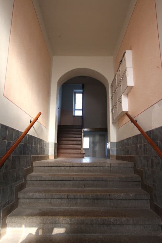 Treppenaufgang mit Steintufen, seitlich dunkle Fliesen und helle Wände, oben Durchgang mit Rundbogen, Holzgeländer, Oberlicht