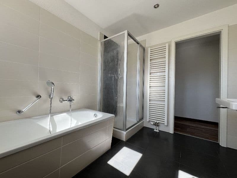 Badezimmer mit weißer Badewanne, Duschkabine mit Glastüren, schwarzem Fliesenboden und heller Wandfliesen