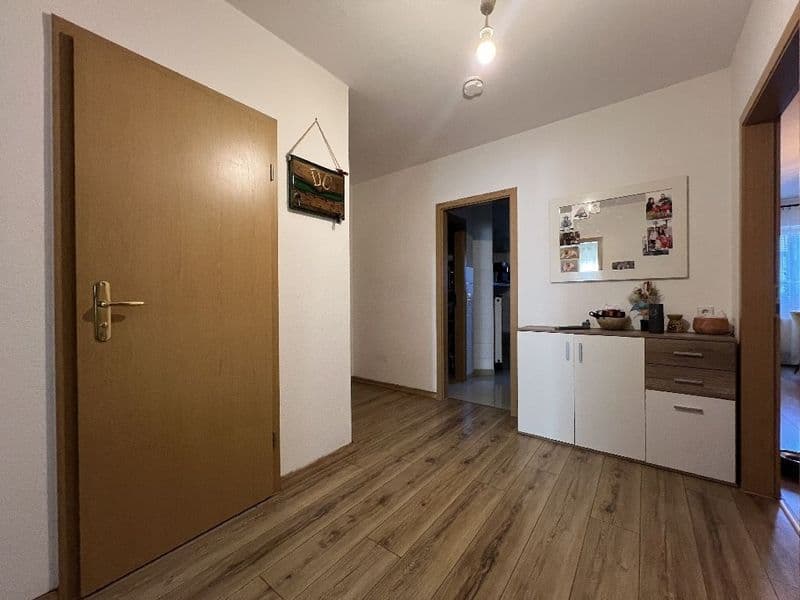 Flur mit Laminatboden, braune Holztür links, weiße Wände, Kommode mit weißen Schranktüren und Spiegel rechts