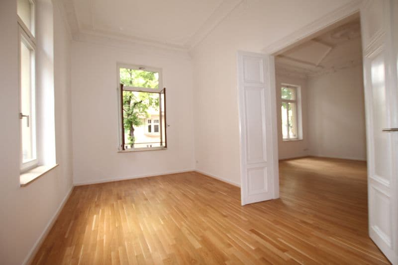 Leerer Raum mit Parkettboden, weißen Wänden, zwei Fenster und offene Türe zu Nebenraum