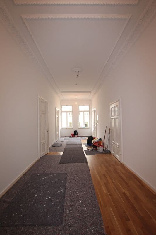 Langer Flur mit Stuckdecke, weiße Türen beidseitig, teilweise Parkettboden und dunkler Teppichbelag, Fenster am Ende