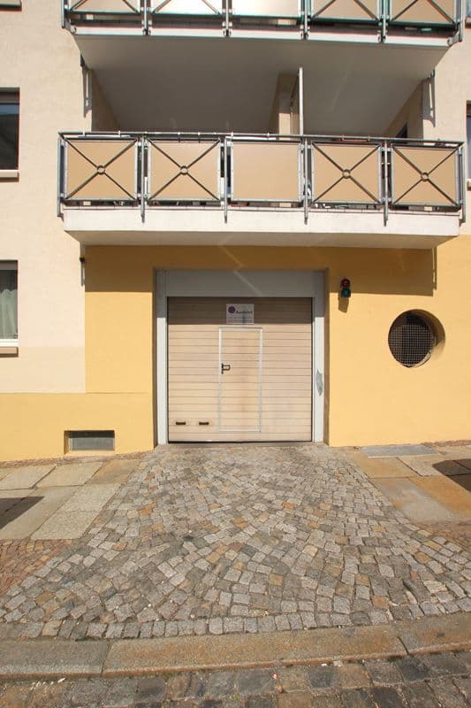 Hausfront mit gelbem Putz, Garagen-Rolltor, zwei Balkonen mit X-Gittern und Kopfsteinpflaster-Zufahrt