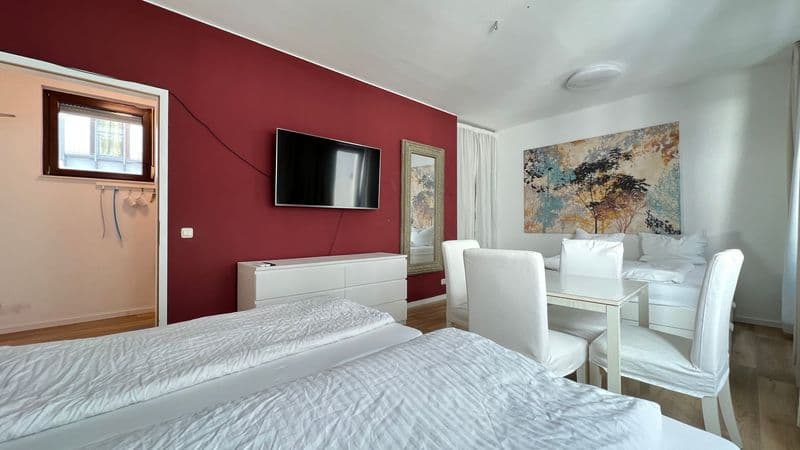 Schlafzimmer mit dunkelroter Wand, Doppelbett, weißer Kommode, TV-Gerät und Essbereich mit weißen Stühlen