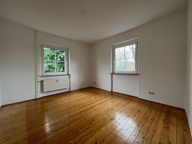 Leeres Zimmer mit Holzdielen, zwei Fenster mit weißem Rahmen, Heizkörper unter linkem Fenster, weiße Wände
