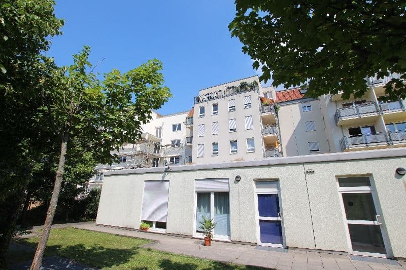 Eingeschossiger Flachbau mit weißer Fassade und Glastüren, dahinter mehrgeschossiges Wohnhaus mit Balkonen