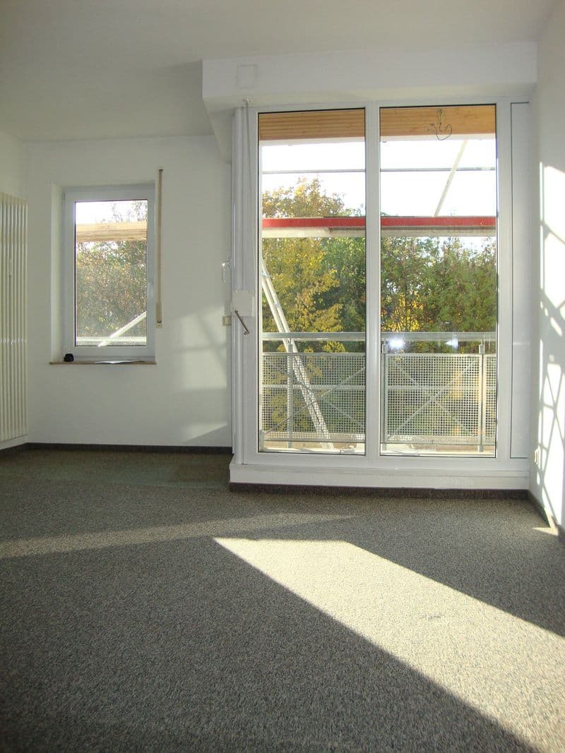 Leeres Wohnzimmer mit Teppichboden, weiße Wände, große Glasfenstertür mit Balkonzugang und Ausblick auf Baumbestand