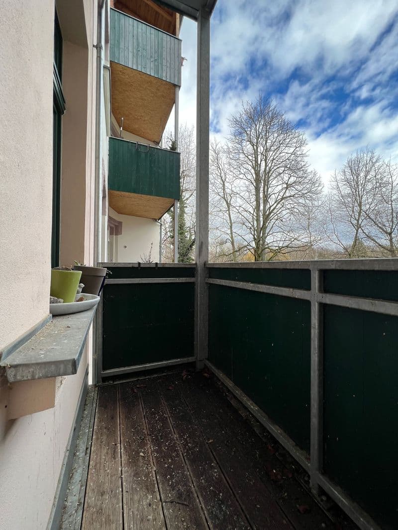 Balkon mit dunklem Holzbelag, grünem Sichtschutz, Blick auf Wohngebäude mit Balkonen und kahle Bäume